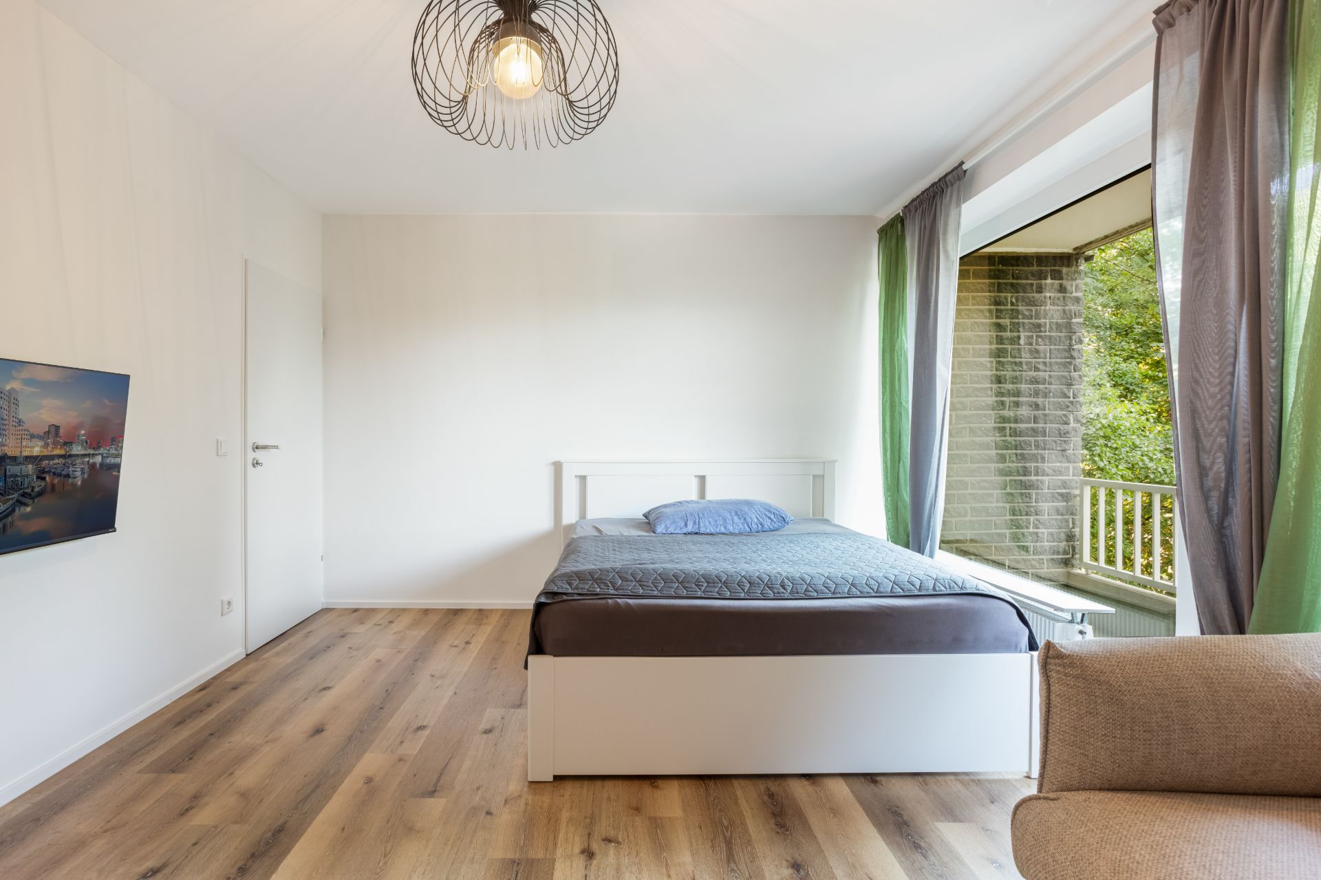 Helles Schlafzimmer mit großem Bett, Holzfußboden, Fernseher und Zugang zu einem Balkon.