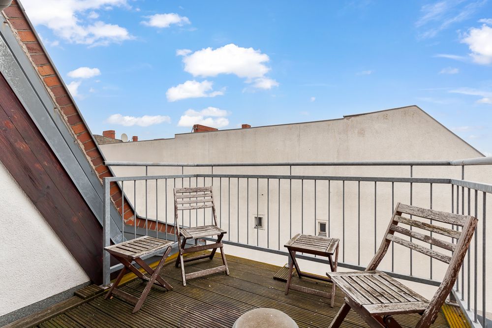 Balkon mit Holzboden, drei Holzstühlen und einem kleinen Tisch, Blick auf Nachbargebäude.