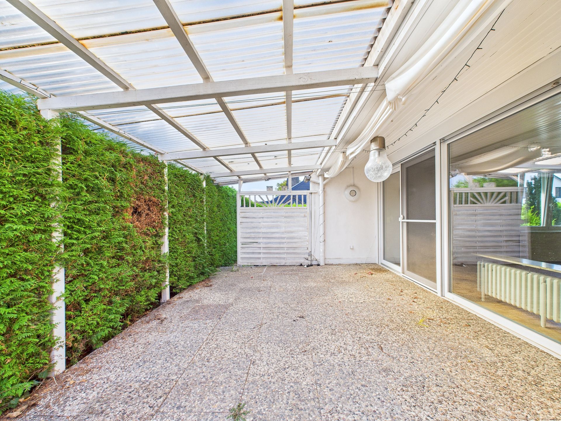Überdachte Terrasse mit Glasdach, Steinboden und grüner Hecke an der Seite.