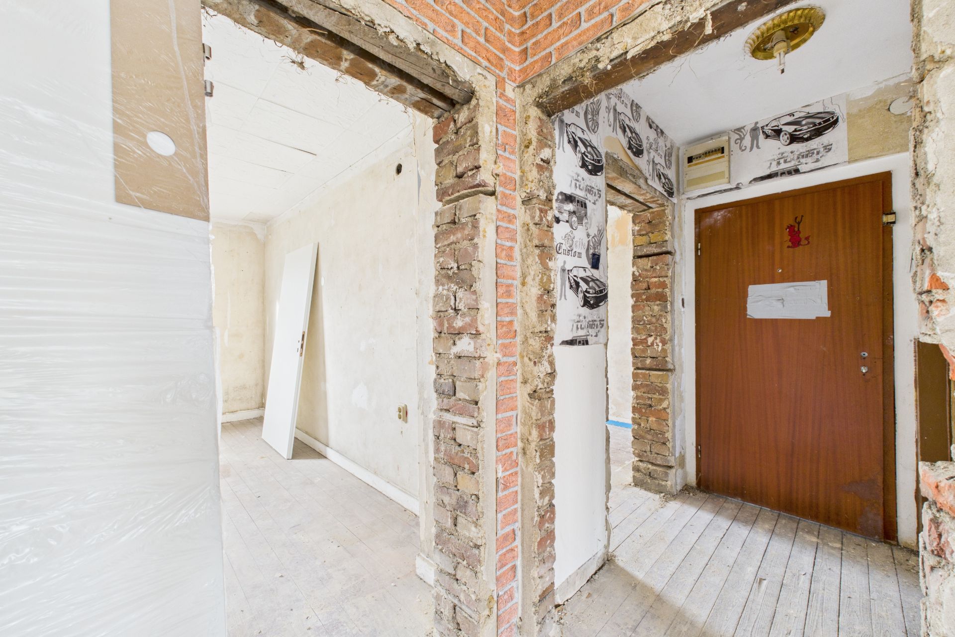 Flur einer renovierungsbedürftigen Wohnung mit sichtbaren Backsteinwänden und offener Holztür.