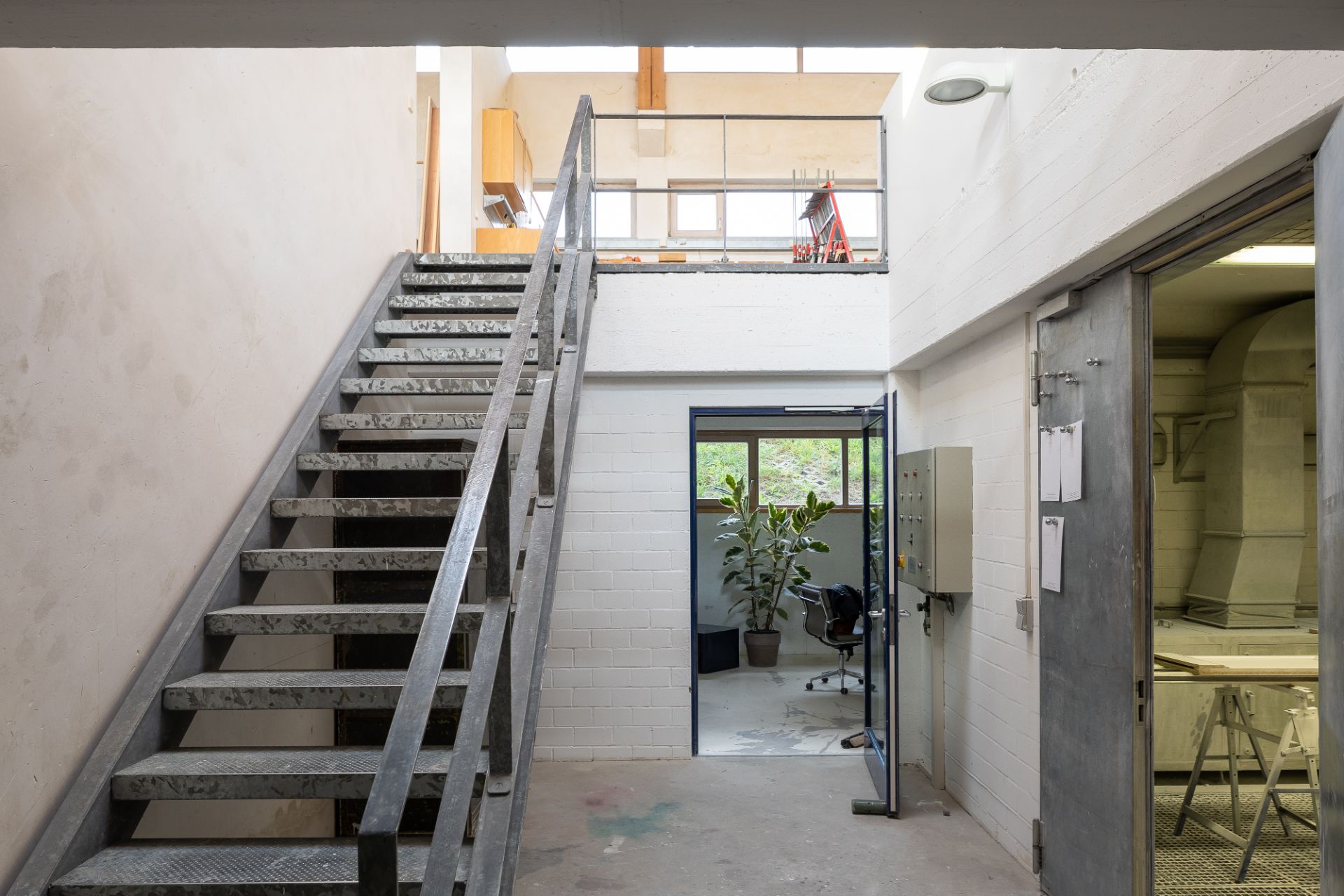 Heller Flur mit Metalltreppe, Glastür zu einem Raum mit Pflanze und Schreibtisch.