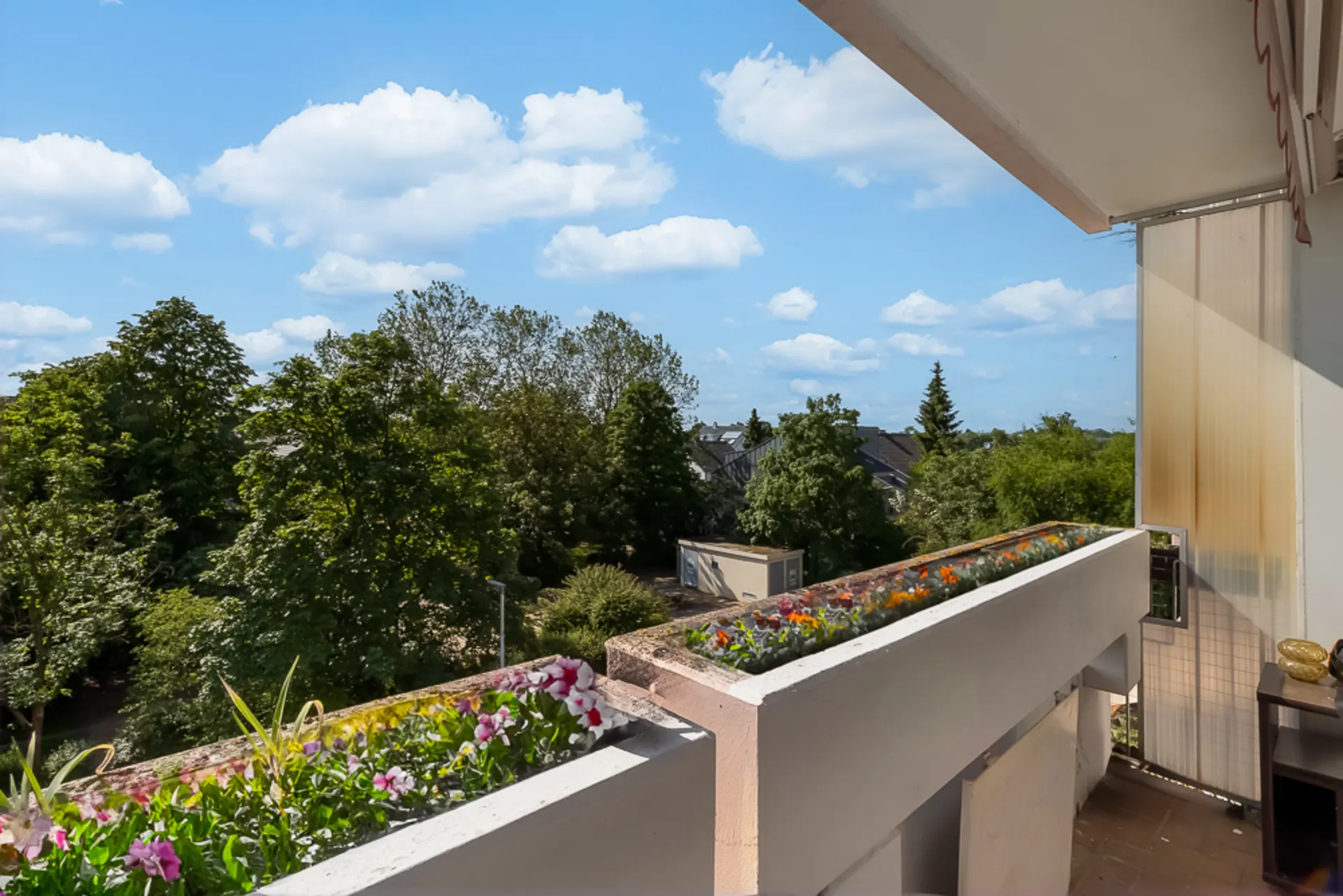 Balkon mit Blumenkästen, Blick auf grüne Bäume und blauen Himmel mit wenigen Wolken.