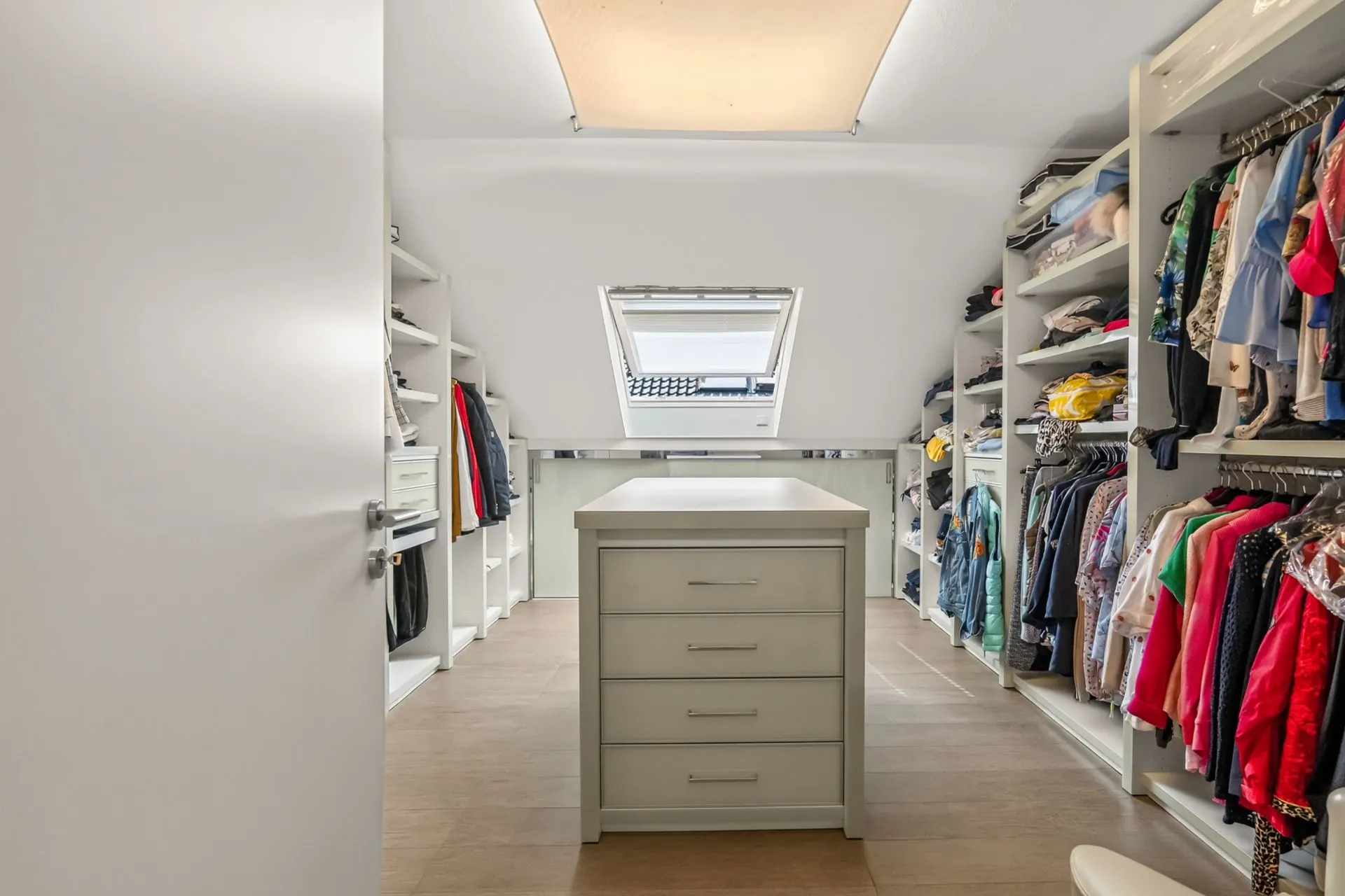 Begehbarer Kleiderschrank mit Regalen, Kleiderstangen und Kommode unter einem Dachfenster.