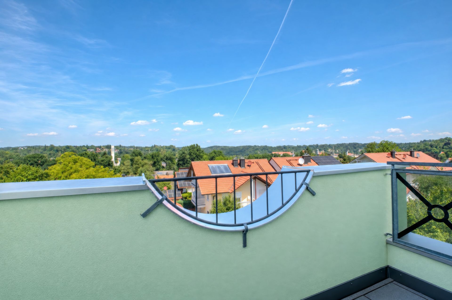 Balkon mit grünem Geländer und Blick auf Dächer und Bäume unter blauem Himmel.