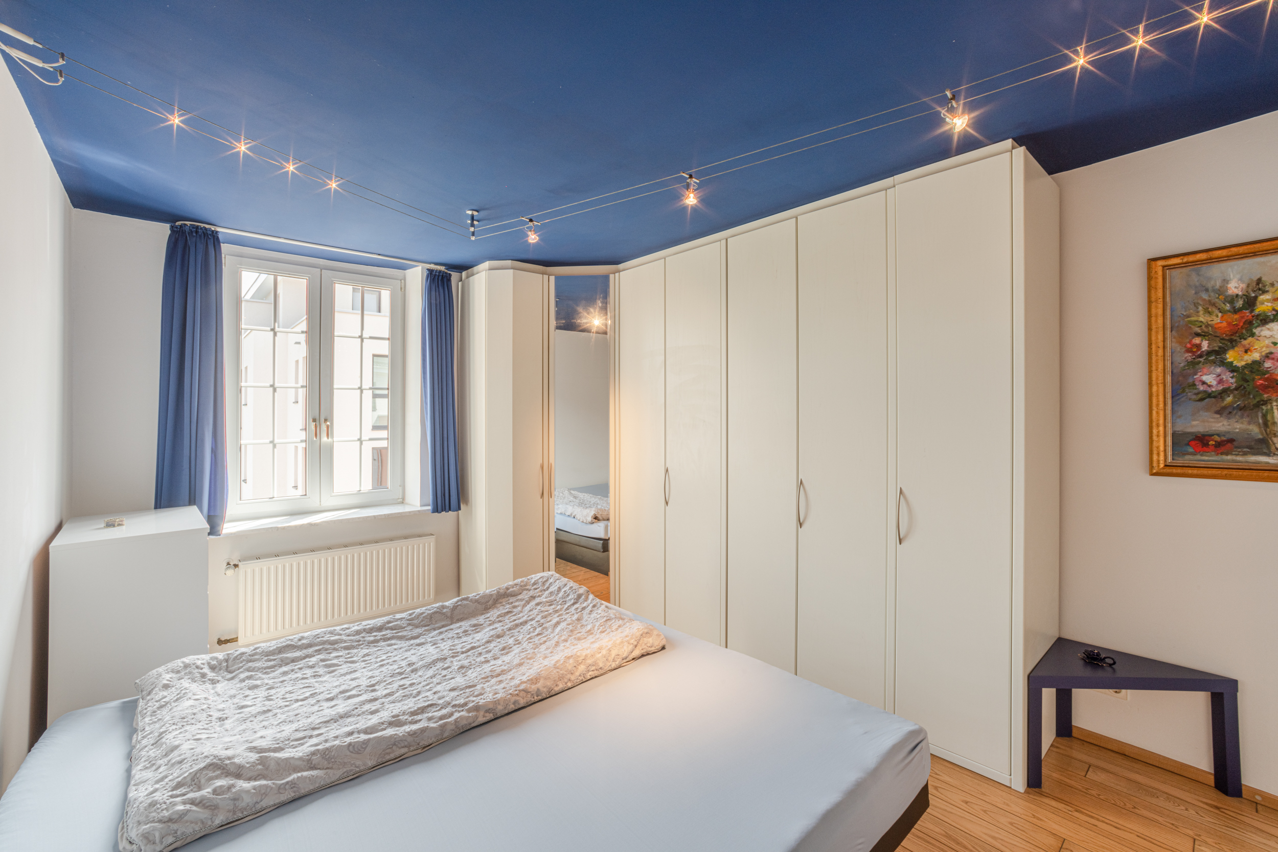 Helles Schlafzimmer mit großem Schrank, blauem Vorhang und Bett unter einer blauen Decke.