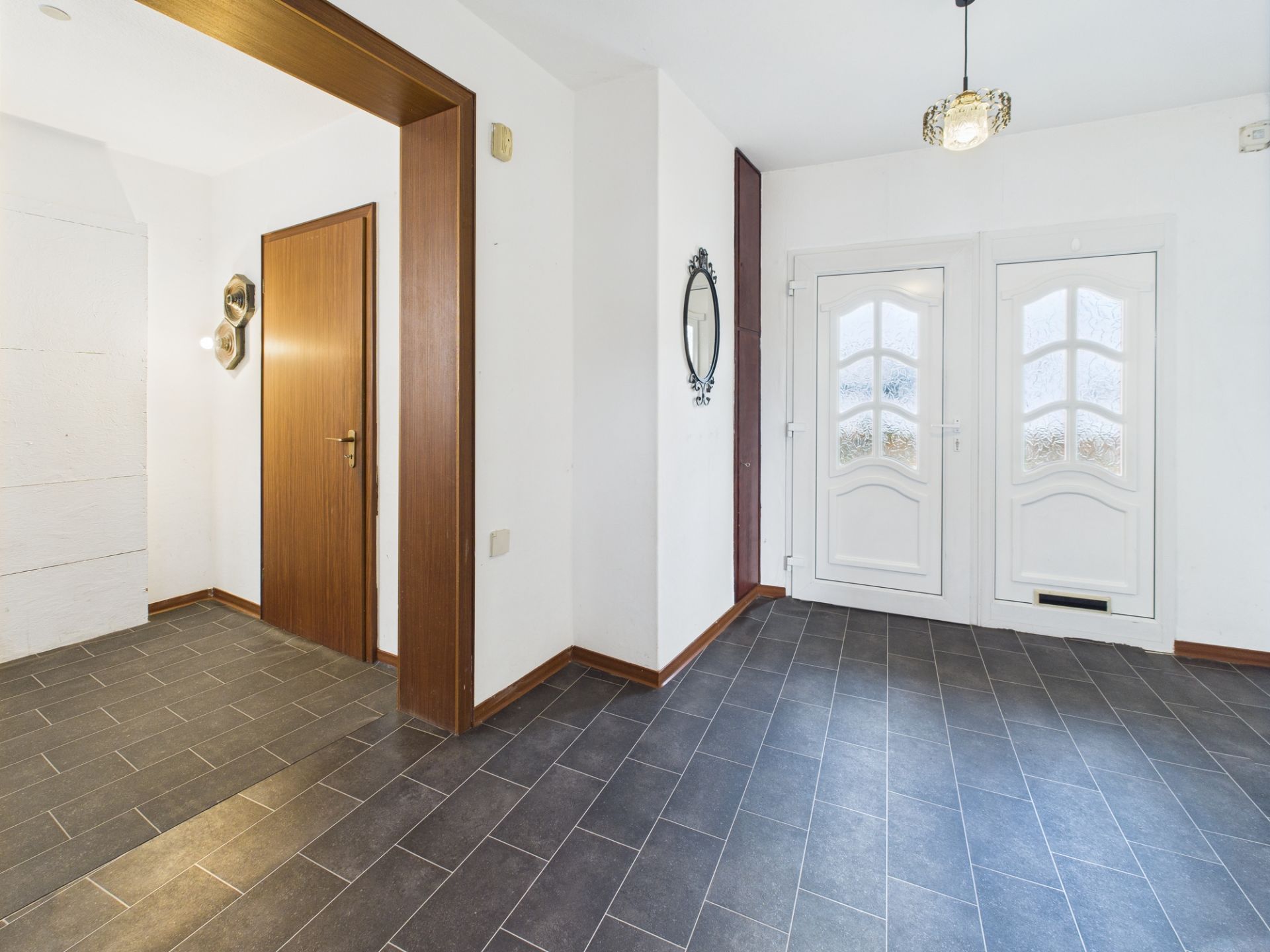 Heller Flur mit weißen Türen, Spiegel und dunklem Fliesenboden in einem Wohnhaus.