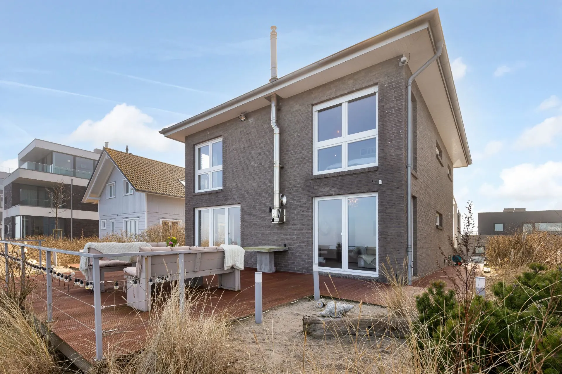 Modernes zweistöckiges Haus mit großer Terrasse und Sitzbereich, umgeben von Sand und Gräsern.