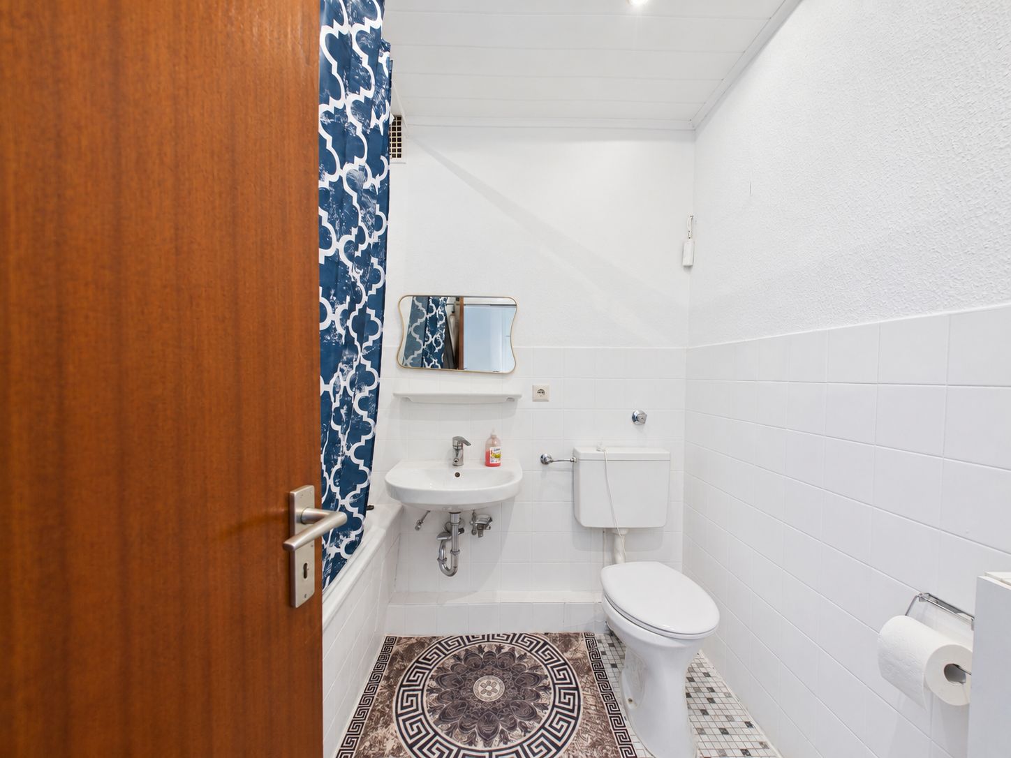 Kleines Badezimmer mit Toilette, Waschbecken, Spiegel und gemustertem Duschvorhang.