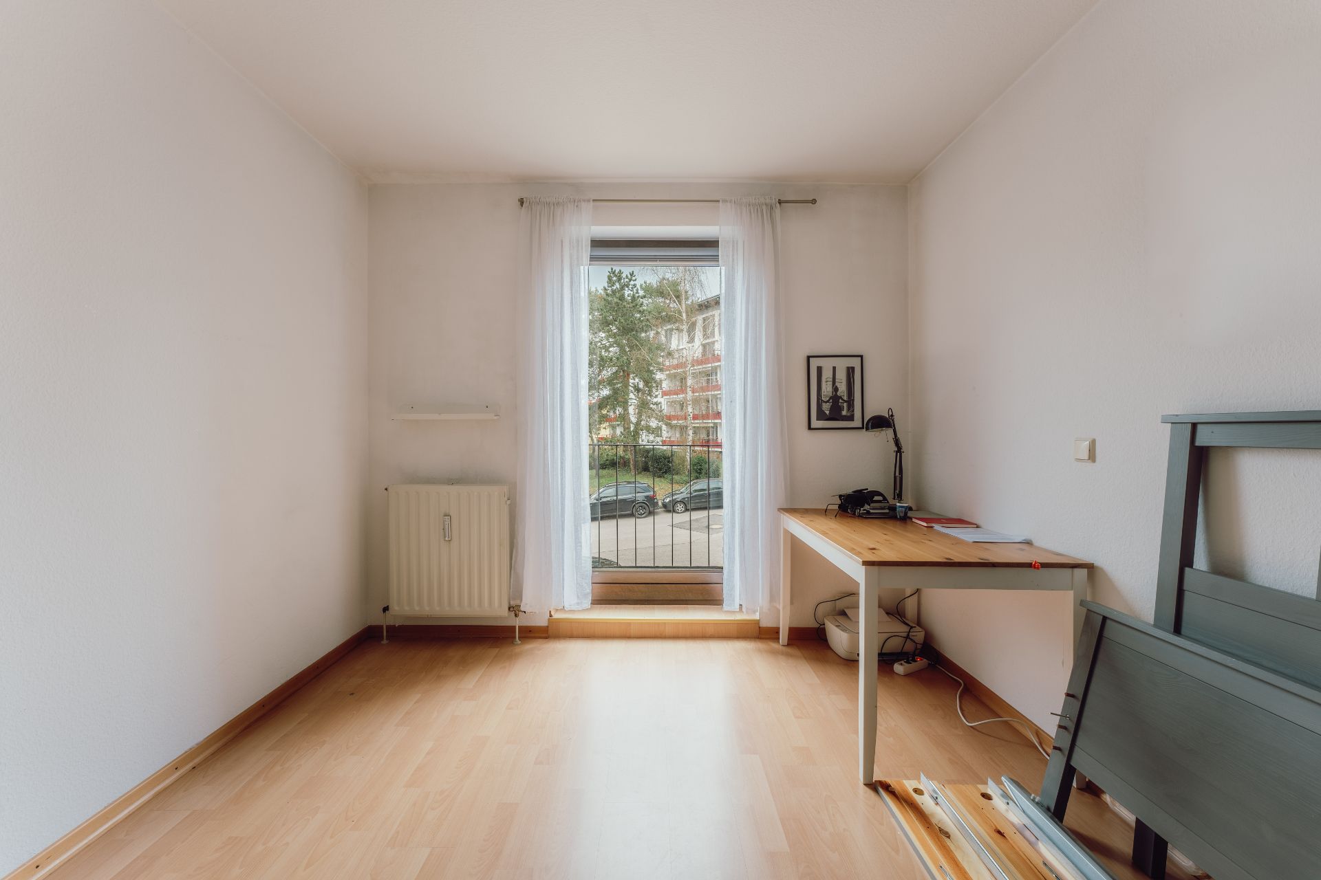 Helles Zimmer mit Holzfußboden, Schreibtisch, Fenster mit Vorhang und Heizkörper.