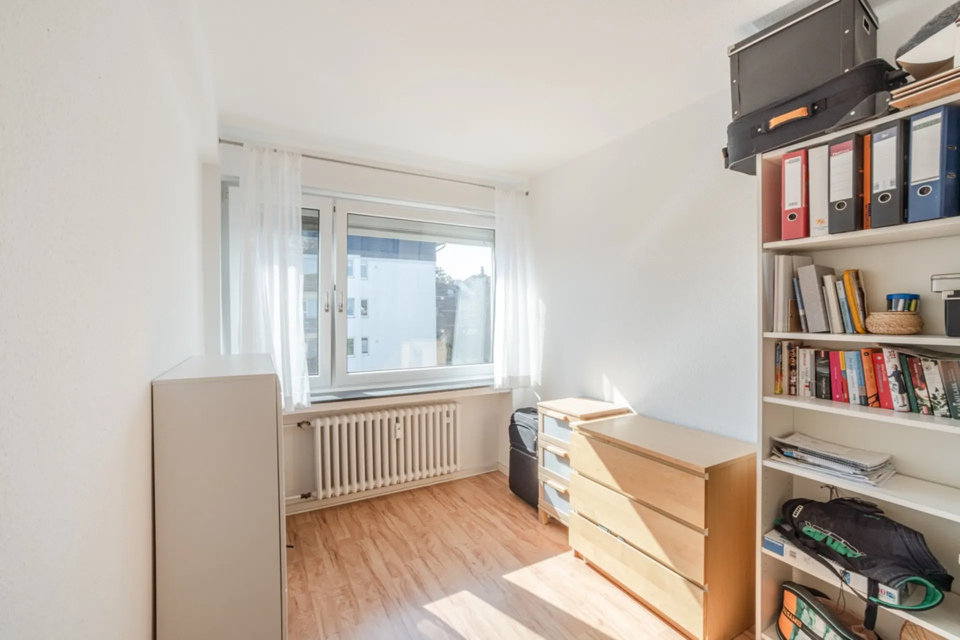 Helles Zimmer mit großem Fenster, Holzfußboden, Regal, Kommode und Aktenschrank.