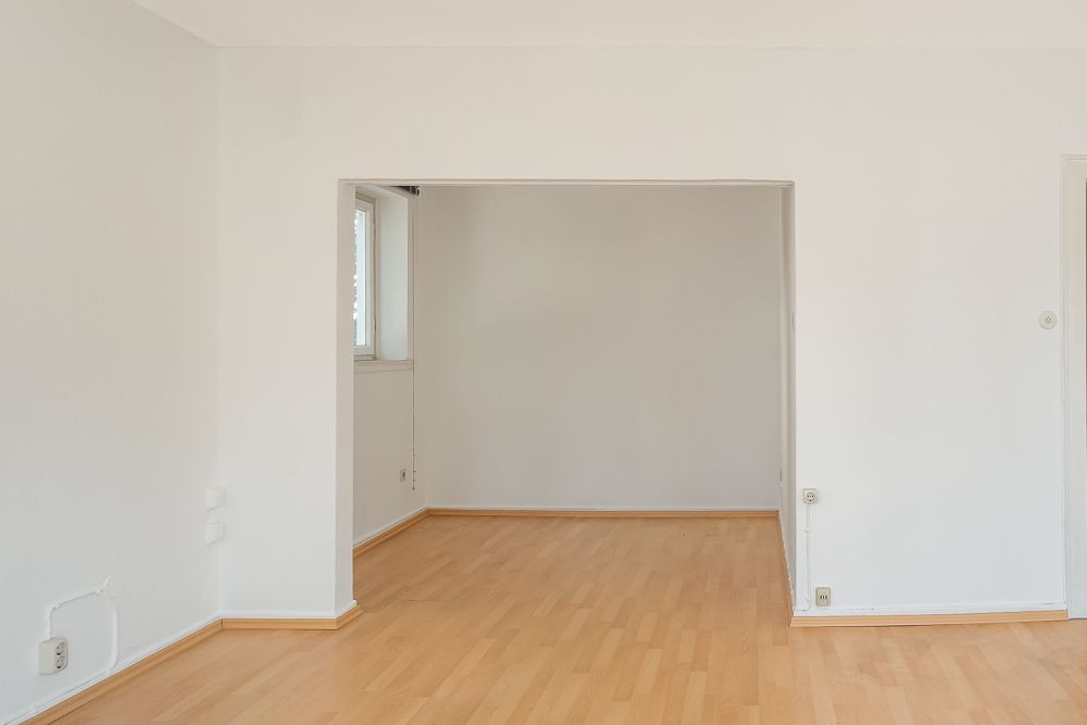 Leerer Raum mit hellem Holzfußboden und weißen Wänden, ein Durchgang führt in ein weiteres Zimmer.