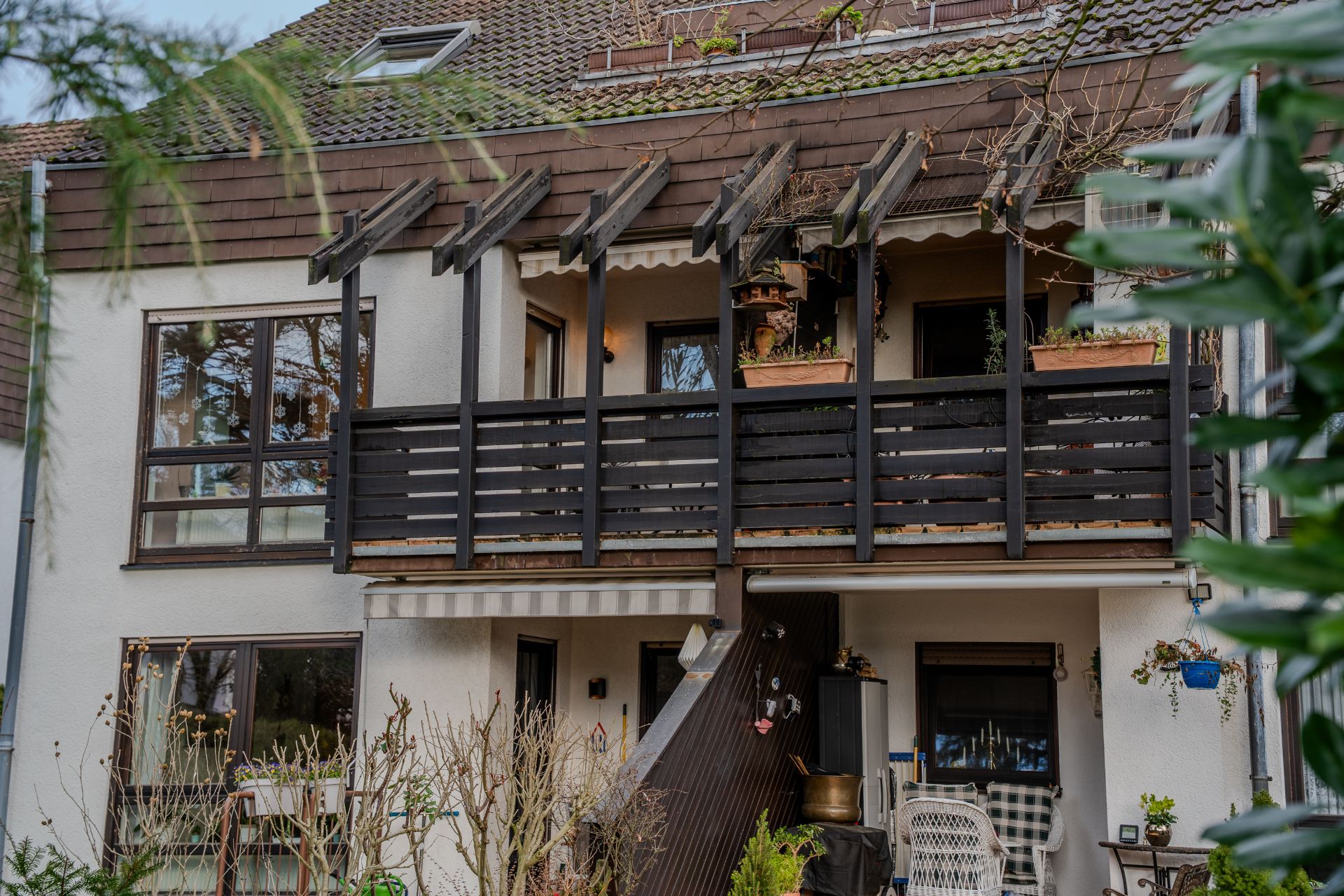 Zweistöckiges Wohnhaus mit Balkon, Pflanzen und Sitzmöbeln im Gartenbereich.