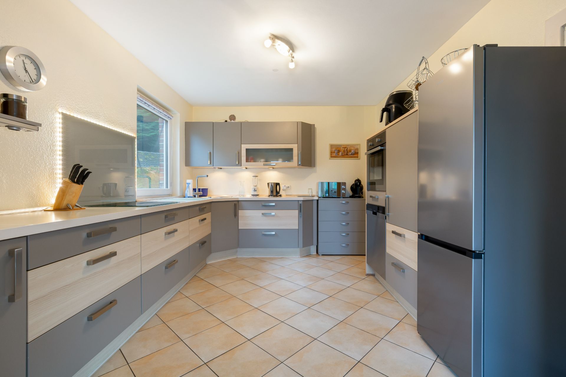 Moderne Küche mit grauen Schränken, großem Kühlschrank, Fenstern und Fliesenboden.