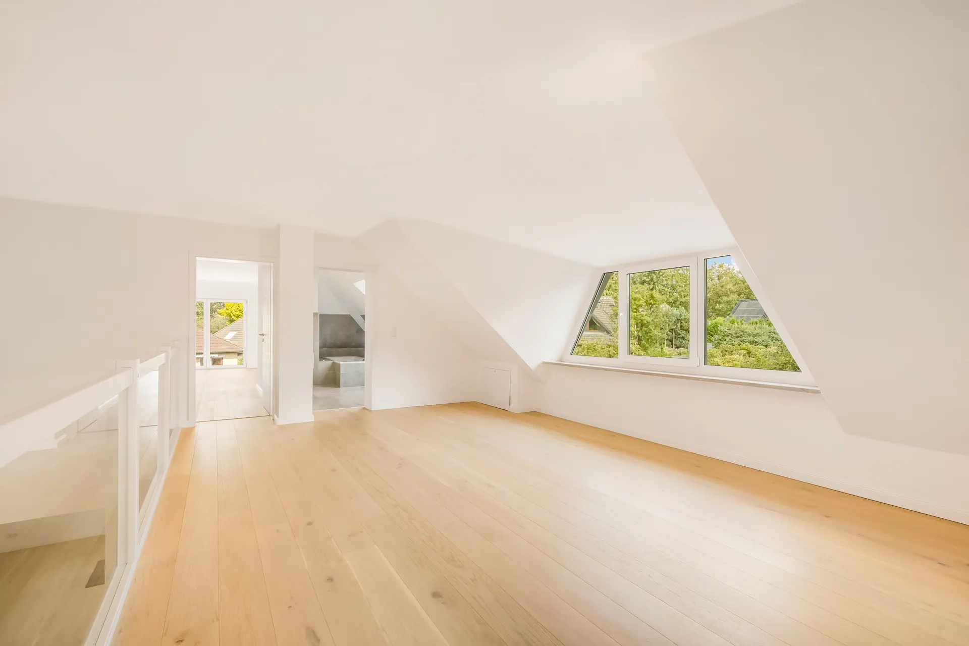 Heller Dachgeschossraum mit Holzfußboden und großen Fenstern mit Blick ins Grüne.