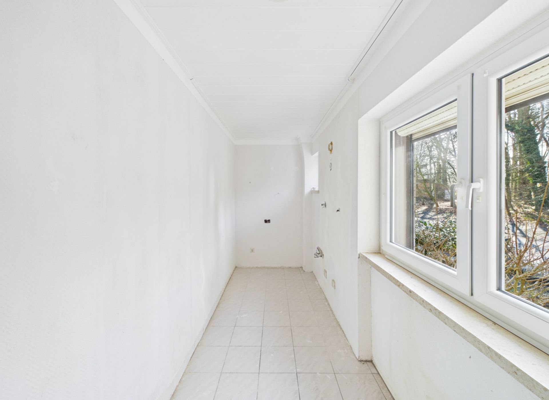 Heller, leerer Raum mit weißen Wänden, Fliesenboden und großem Fenster.