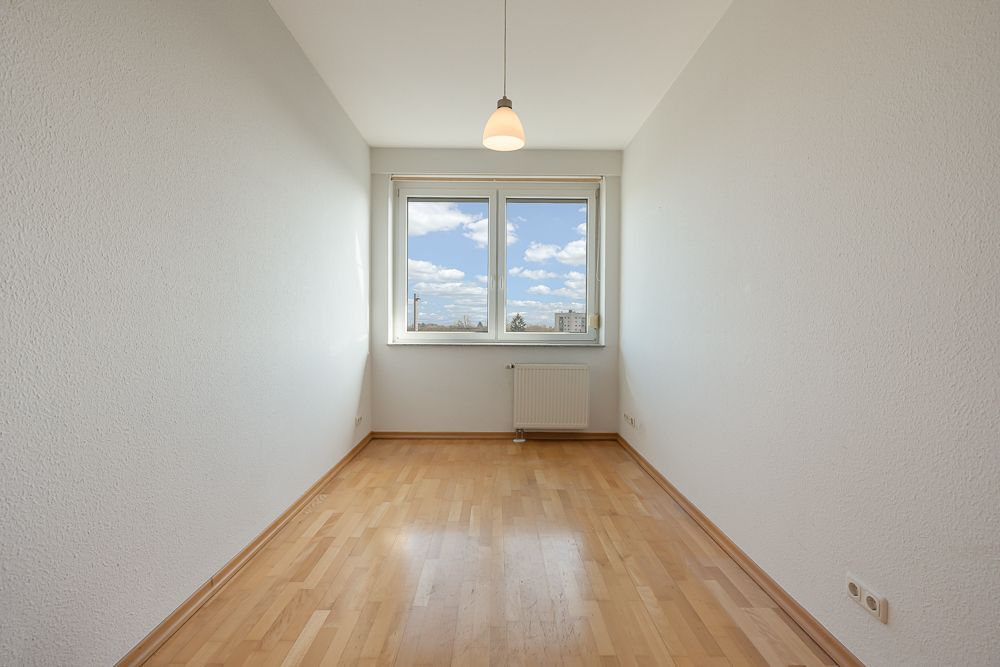 Leerer Raum mit Holzfußboden, weißer Wand, Fenster, Heizung und Deckenlampe.