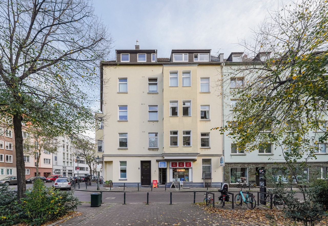Mehrstöckiges Wohnhaus mit Laden im Erdgeschoss, davor Bäume und Fahrräder am Straßenrand.