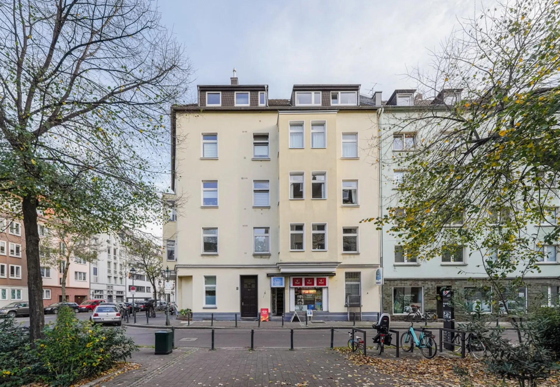 Mehrstöckiges Wohnhaus mit Laden im Erdgeschoss, davor Bäume und Fahrräder am Straßenrand.
