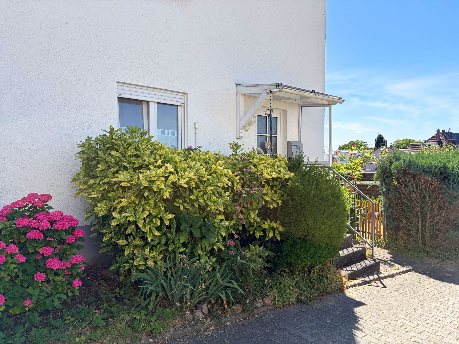 Haus mit weißer Fassade, Fenster, kleiner überdachter Treppe und Sträuchern im Vorgarten.
