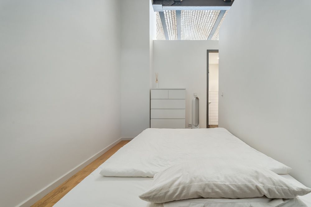 Schlichtes Schlafzimmer mit Bett, weißer Kommode und Holzboden, Licht von Oberlicht.