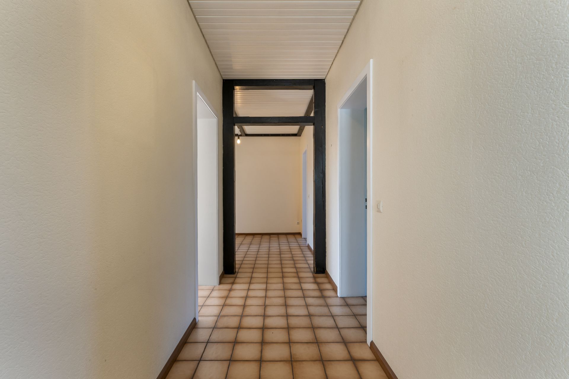 Flur mit Fliesenboden und Türen zu beiden Seiten, helle Wände, sichtbare schwarze Balken.