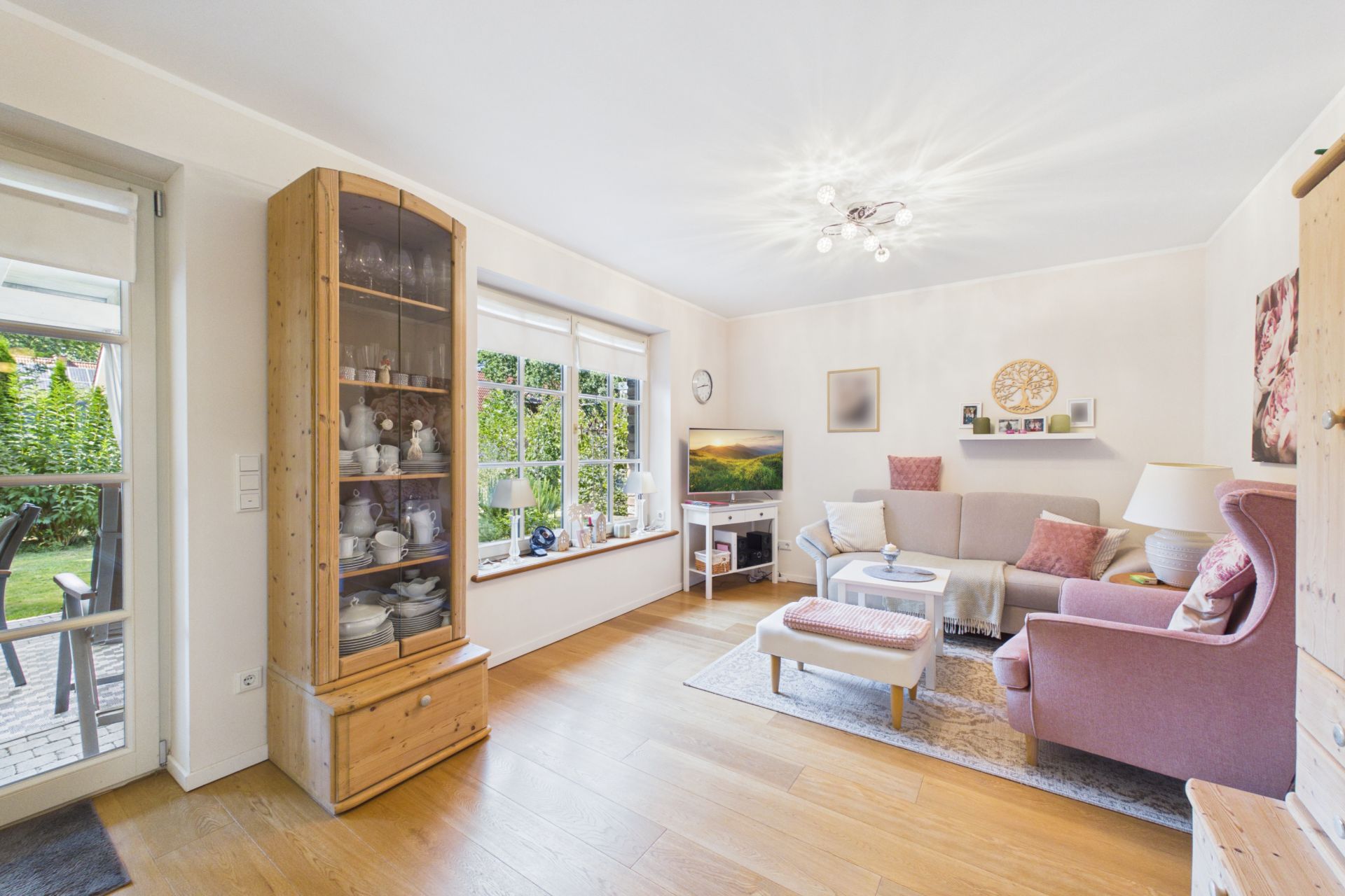 Helles Wohnzimmer mit großem Fenster, Ecksofa, Sessel, Vitrine und Zugang zur Terrasse.