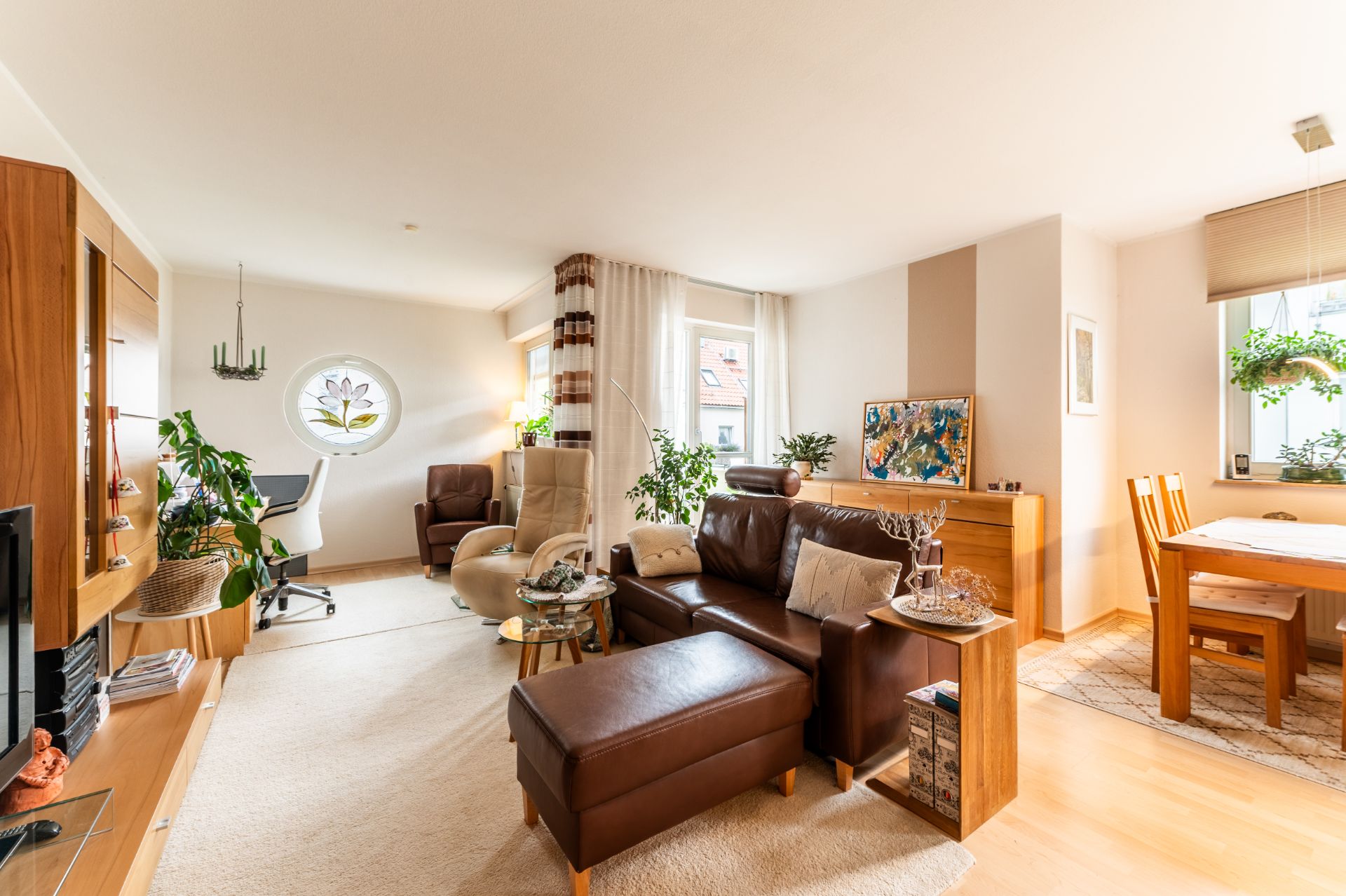 Helles Wohnzimmer mit branem Sofa, Sesseln, Esstisch und vielen Pflanzen.