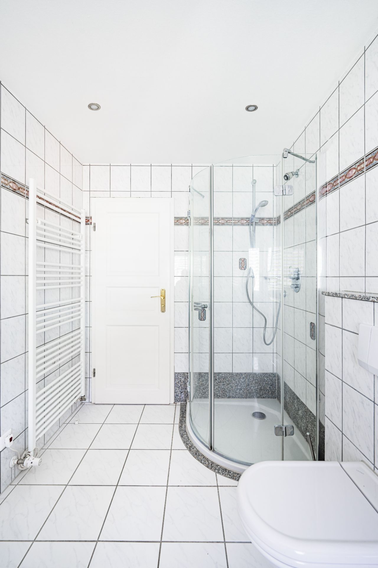 Helles Badezimmer mit Dusche, weiß gekachelten Wänden, Toilette und Handtuchheizkörper.
