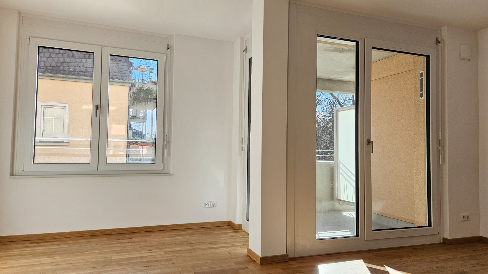 Helles Zimmer mit Parkettboden, zwei Fenstern und einer großen Glastür zu einem Balkon.