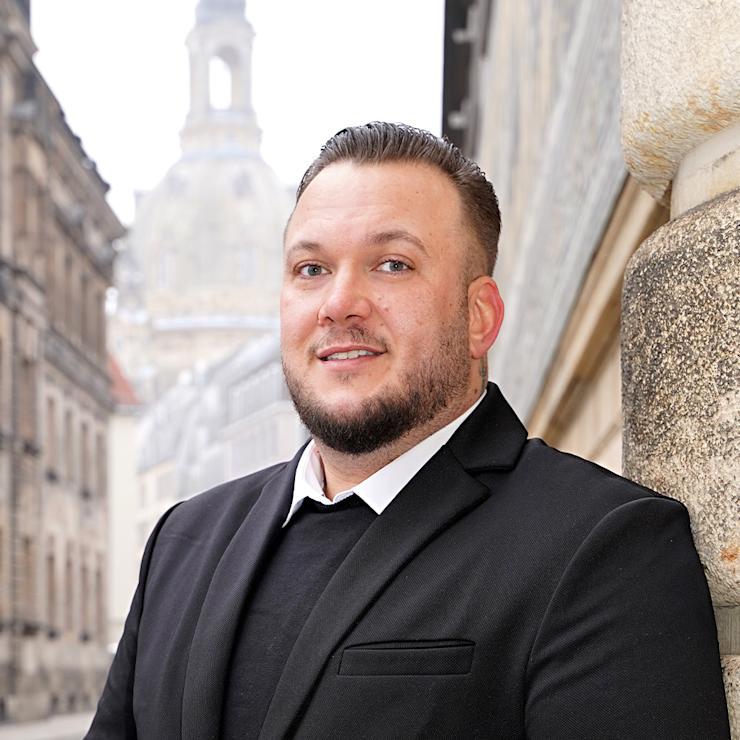 Ein Mann im schwarzen Anzug steht an einer Hauswand in einer Altstadtstraße.