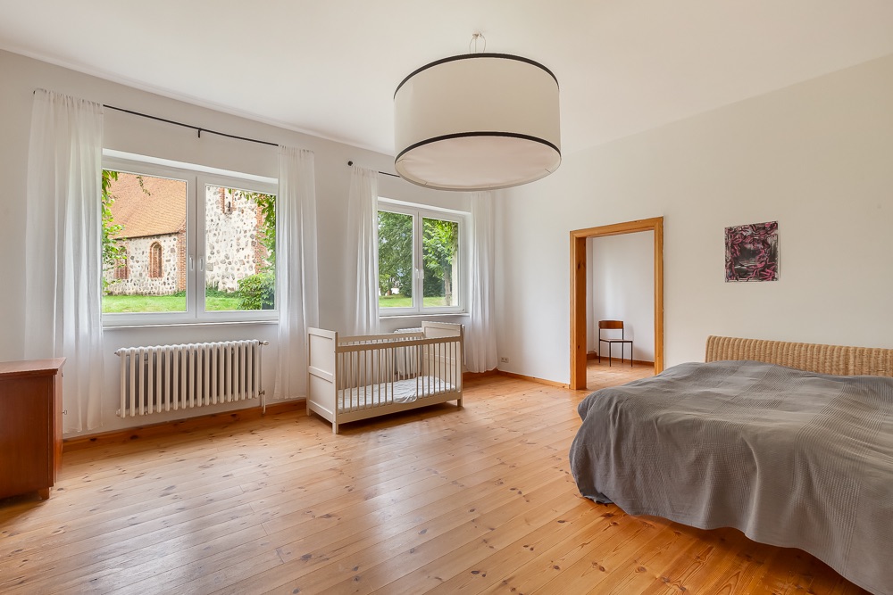 Helles Schlafzimmer mit Babybett, Doppelbett, Holzfußboden und großen Fenstern mit Blick nach draußen.