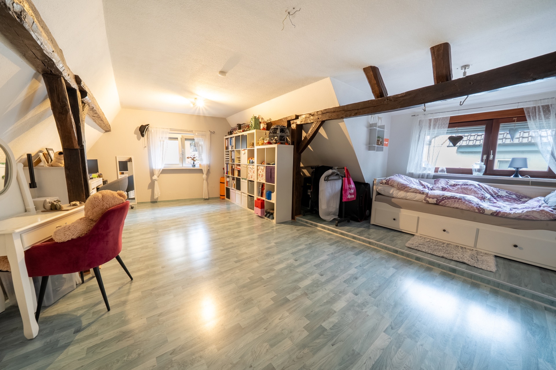 Helles Dachzimmer mit Bett, Regal, Schminktisch und sichtbaren Holzbalken.