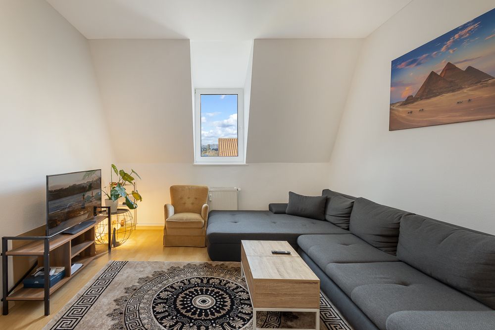 Wohnzimmer mit grauem Ecksofa, Sessel, Fernseher, Teppich und Fenster mit Blick nach draußen.