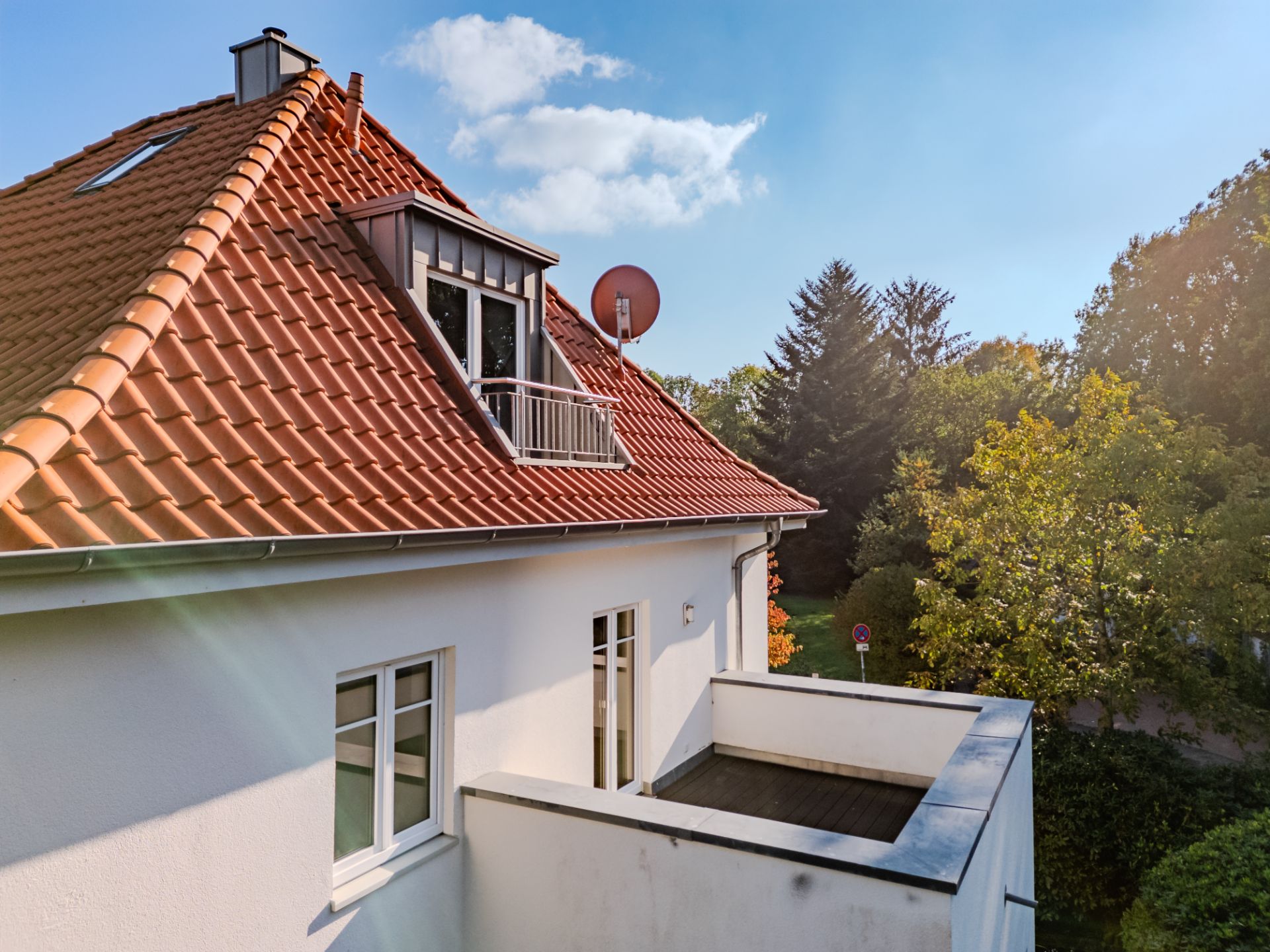 Haus mit rotem Satteldach, Balkon und weißen Wänden, umgeben von Bäumen im Sonnenlicht.