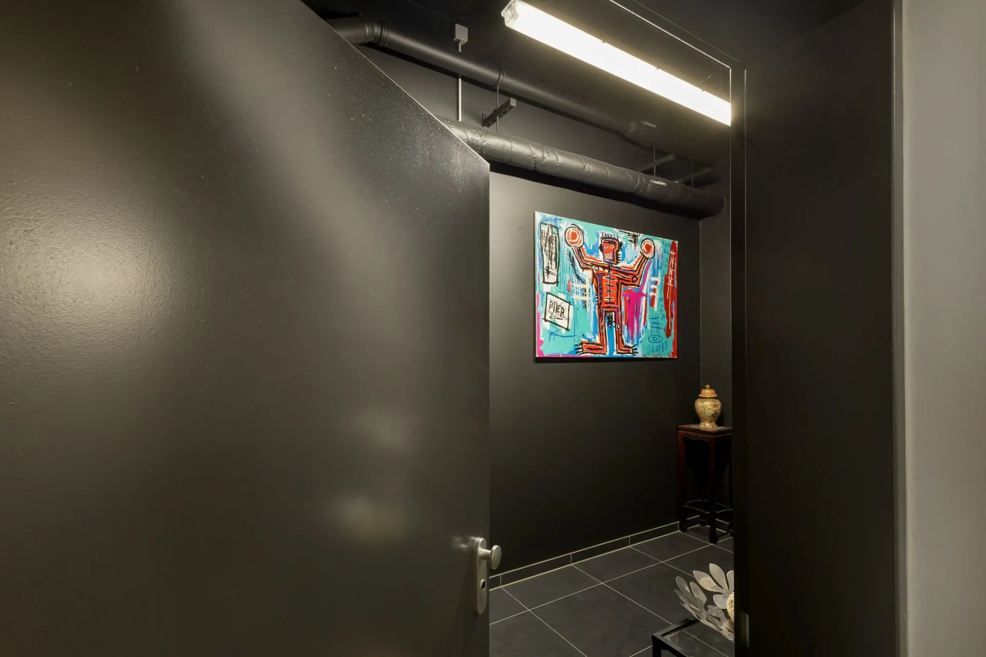 Blick in einen dunklen Raum mit bunter Wandkunst, Vase auf Tisch und sichtbaren Rohren an der Decke.