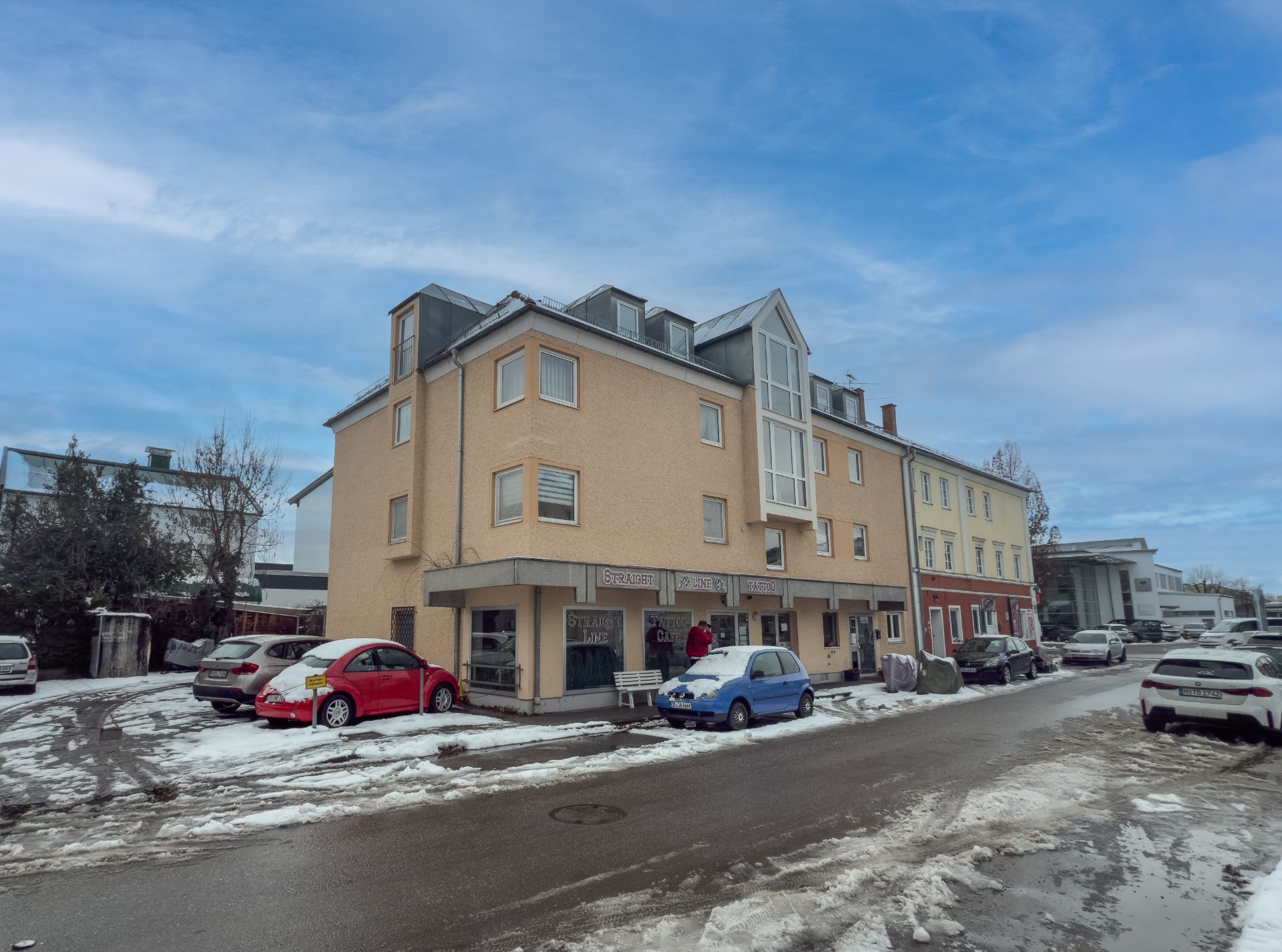 Mehrstöckiges Wohnhaus mit geparkten Autos und Schnee auf der Straße bei Tageslicht.