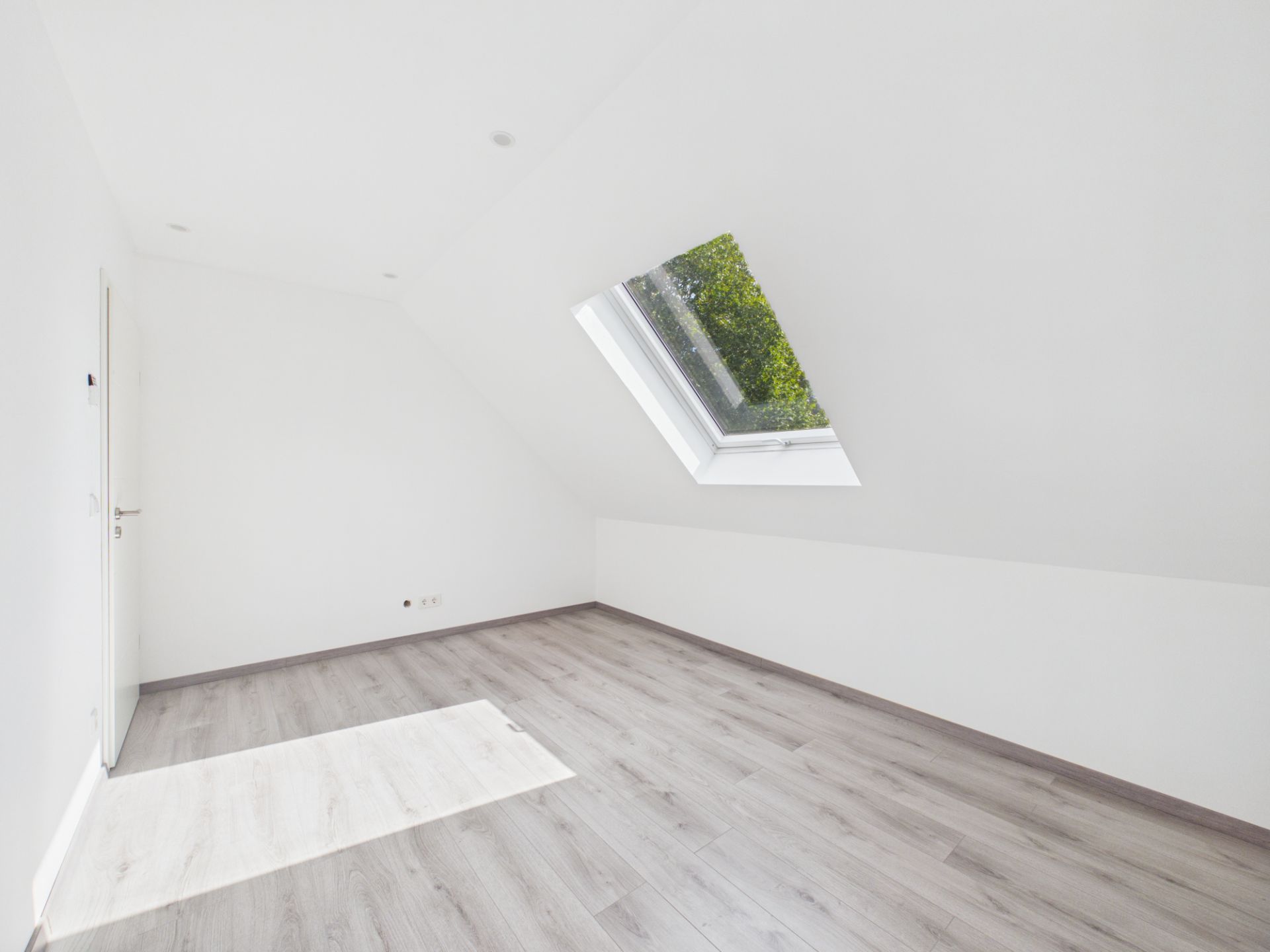 Leeres Zimmer mit schräger Decke, großem Dachfenster und hellem Holzboden.