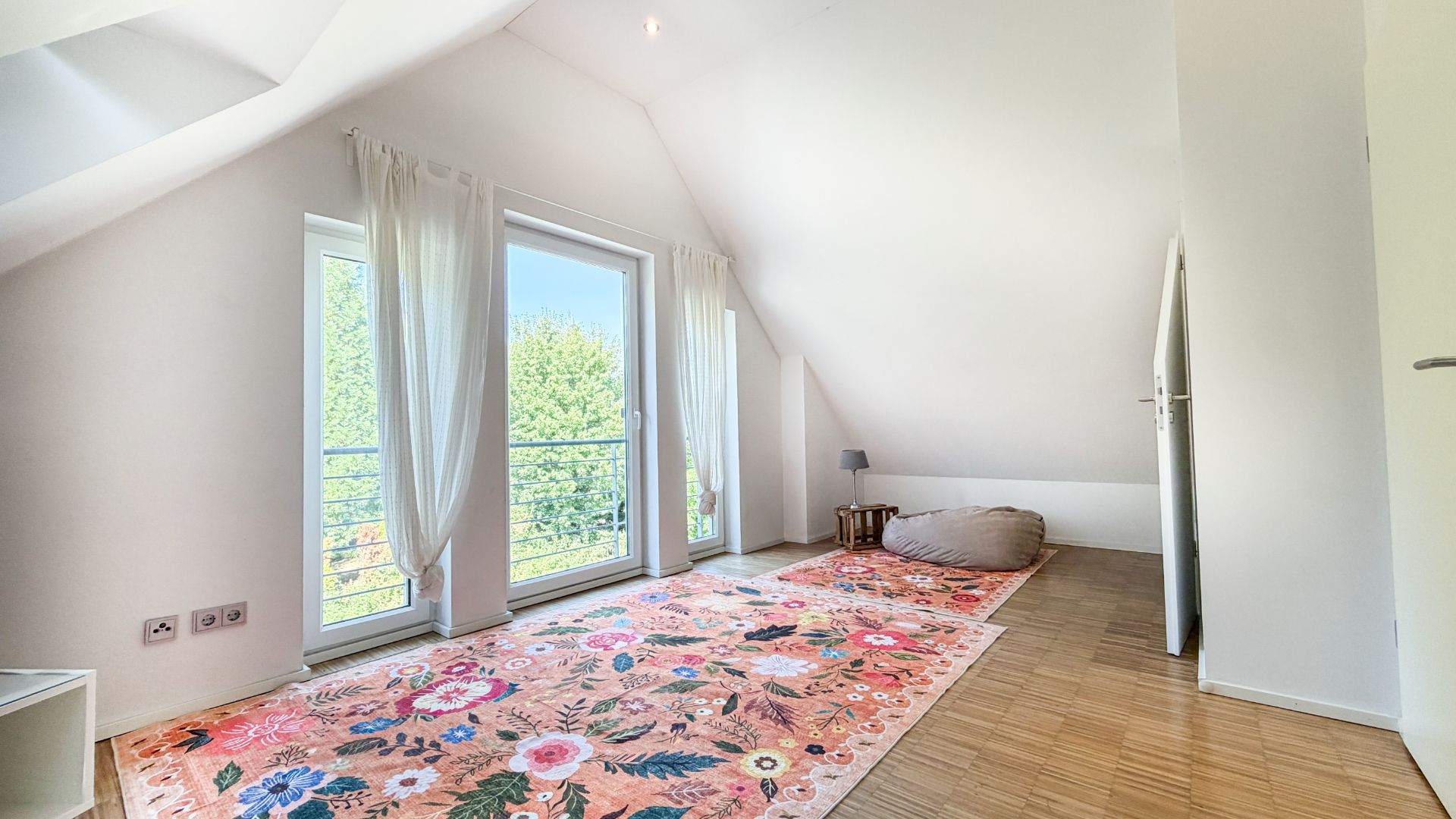 Helles Zimmer mit Dachschräge, buntem Teppich, Sitzsack, kleinem Tisch und Fenstertüren zum Balkon.