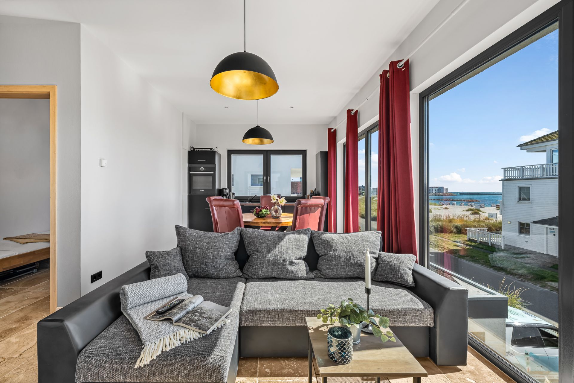 Helles Wohnzimmer mit grauem Sofa, großen Fenstern, Esstisch und Blick aufs Meer.