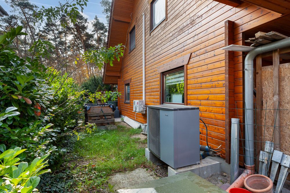 Holzhaus mit Wärmepumpe und Garten, daneben Hecken und Bäume.