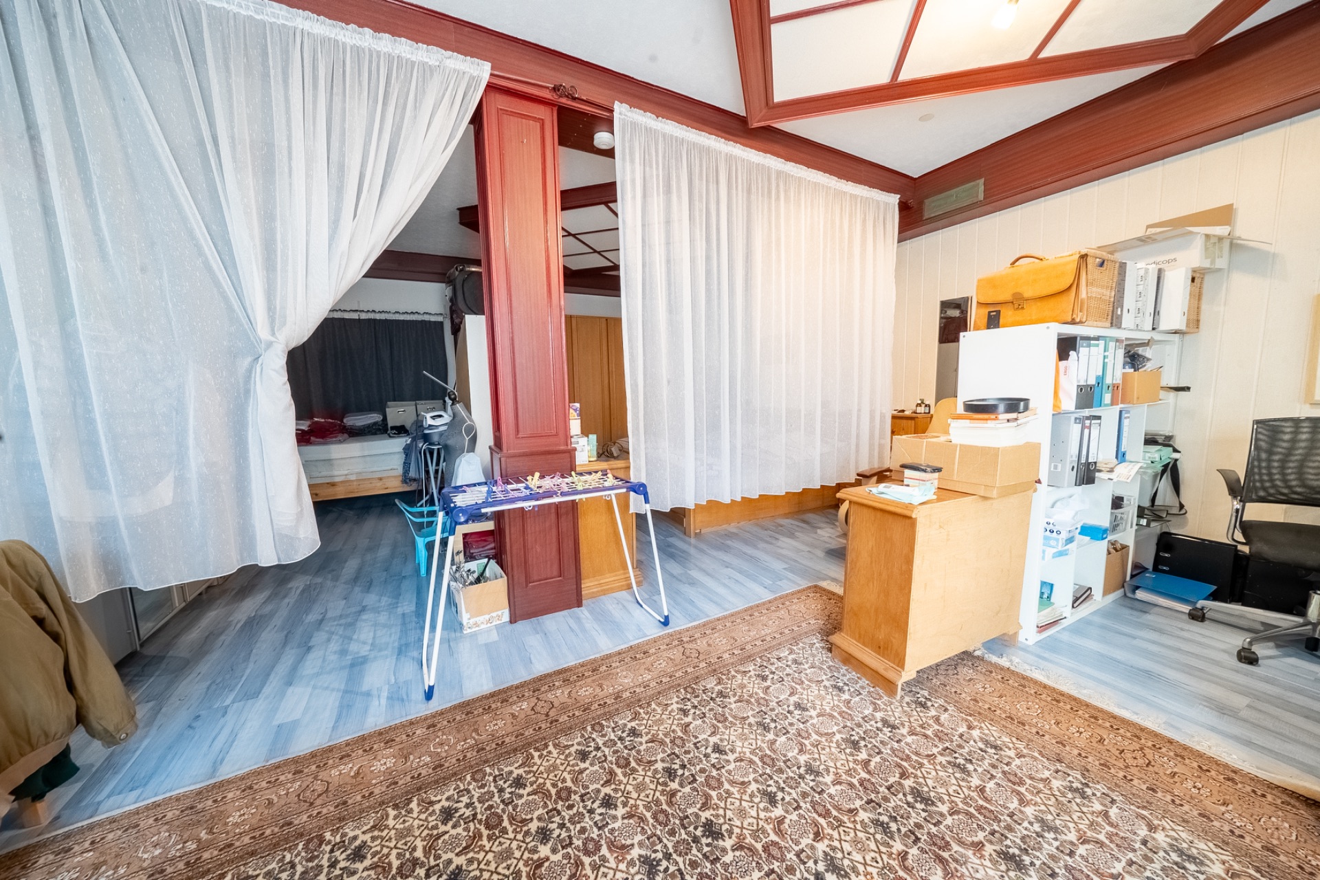 Wohnraum mit Vorhängen, Schreibtisch, Regal und sichtbarem Bett im Hintergrund.