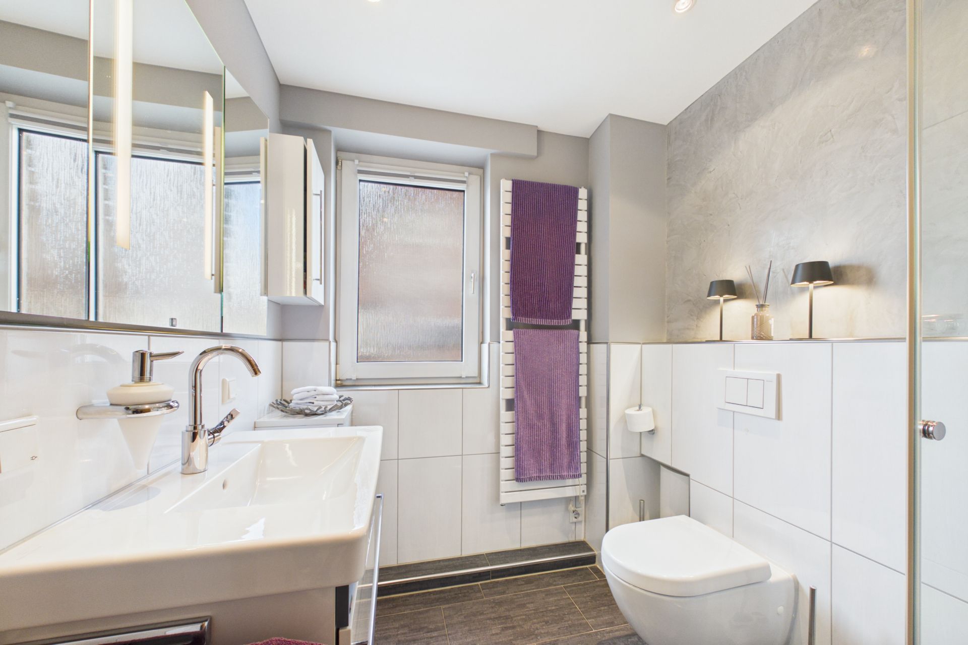 Modernes Badezimmer mit großem Spiegel, Waschbecken, Toilette und lila Handtüchern am Heizkörper.