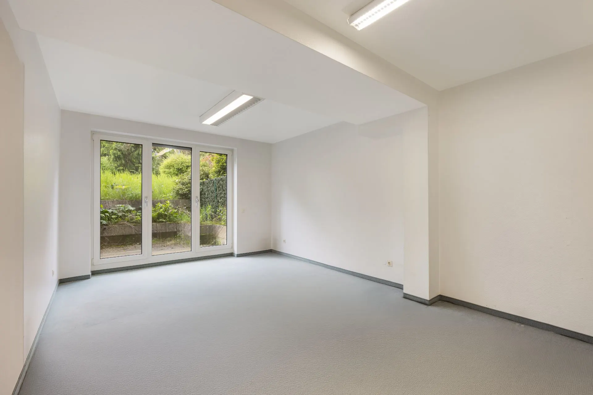 Leerer Raum mit großen Fenstern und Blick auf einen Garten, helle Wände und grauer Boden.