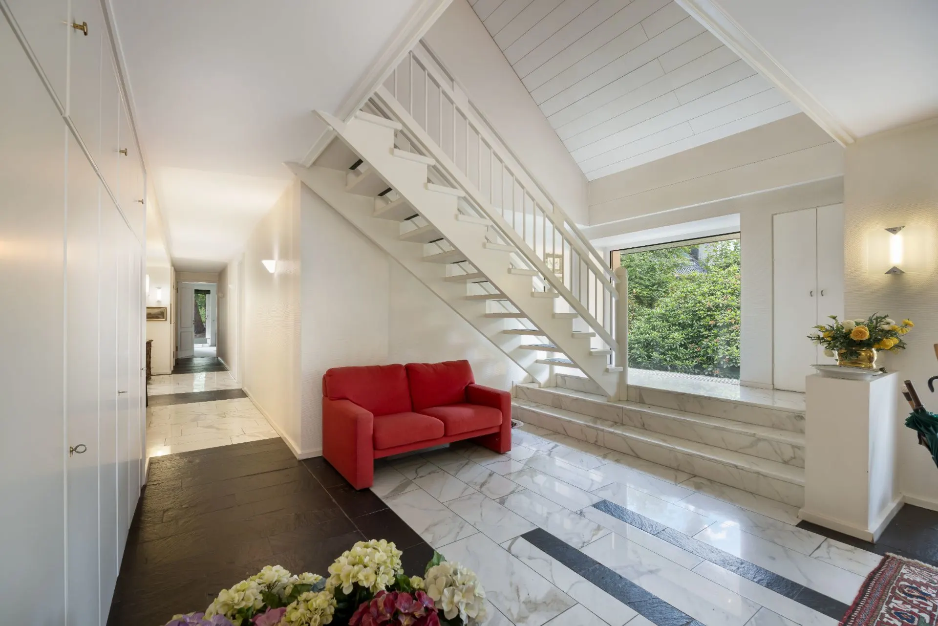 Heller Flur mit Marmorboden, rotem Sofa und Treppe, großes Fenster mit Blick ins Grüne.