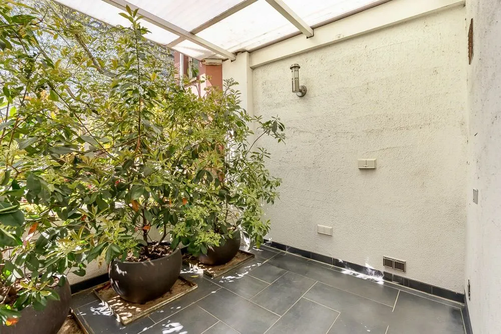 Kleiner, überdachter Balkon mit Fliesenboden und mehreren großen Pflanzen in Töpfen.