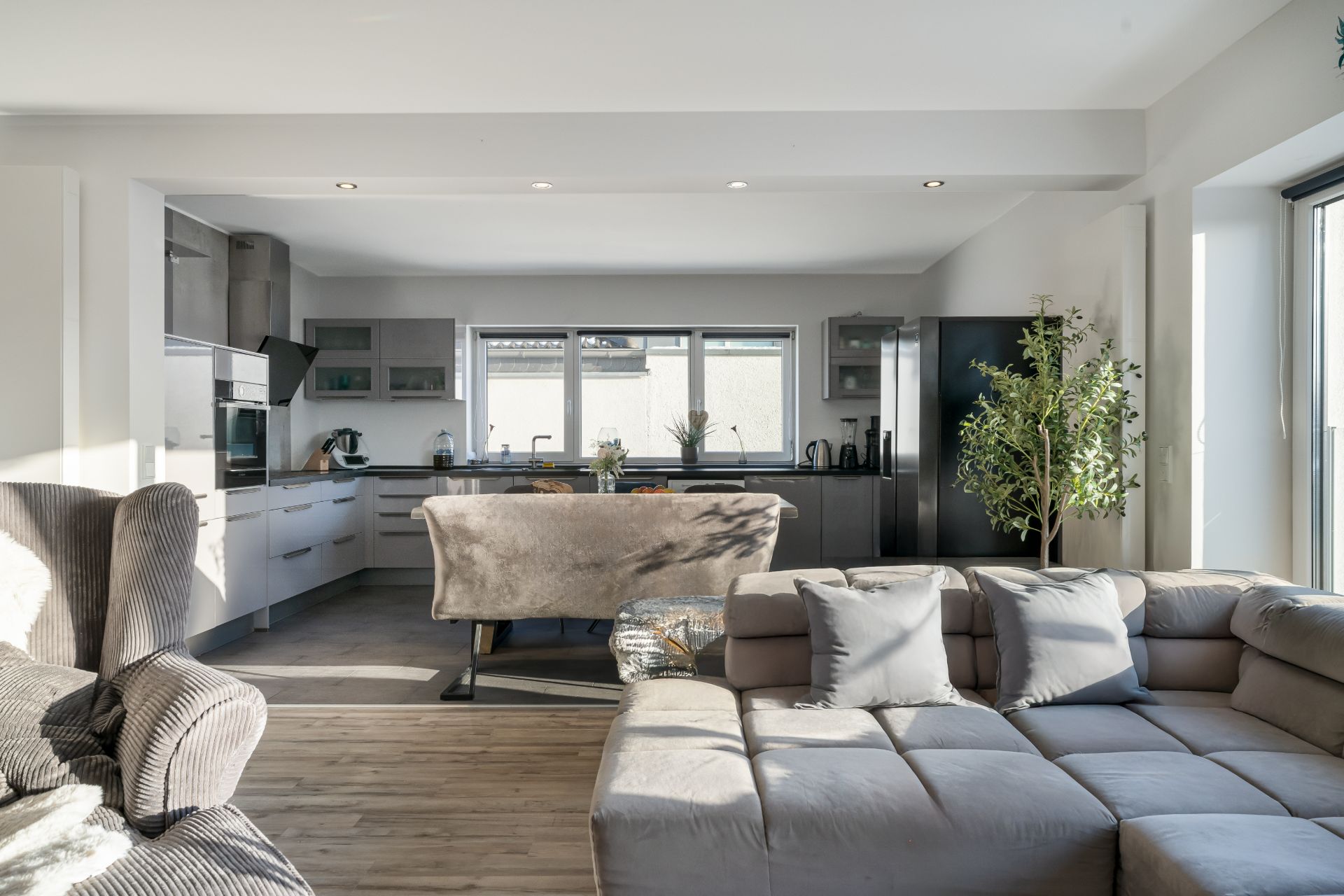 Helles Wohnzimmer mit offener Küche, grauem Sofa, Sessel und Pflanzen, große Fenster im Hintergrund.