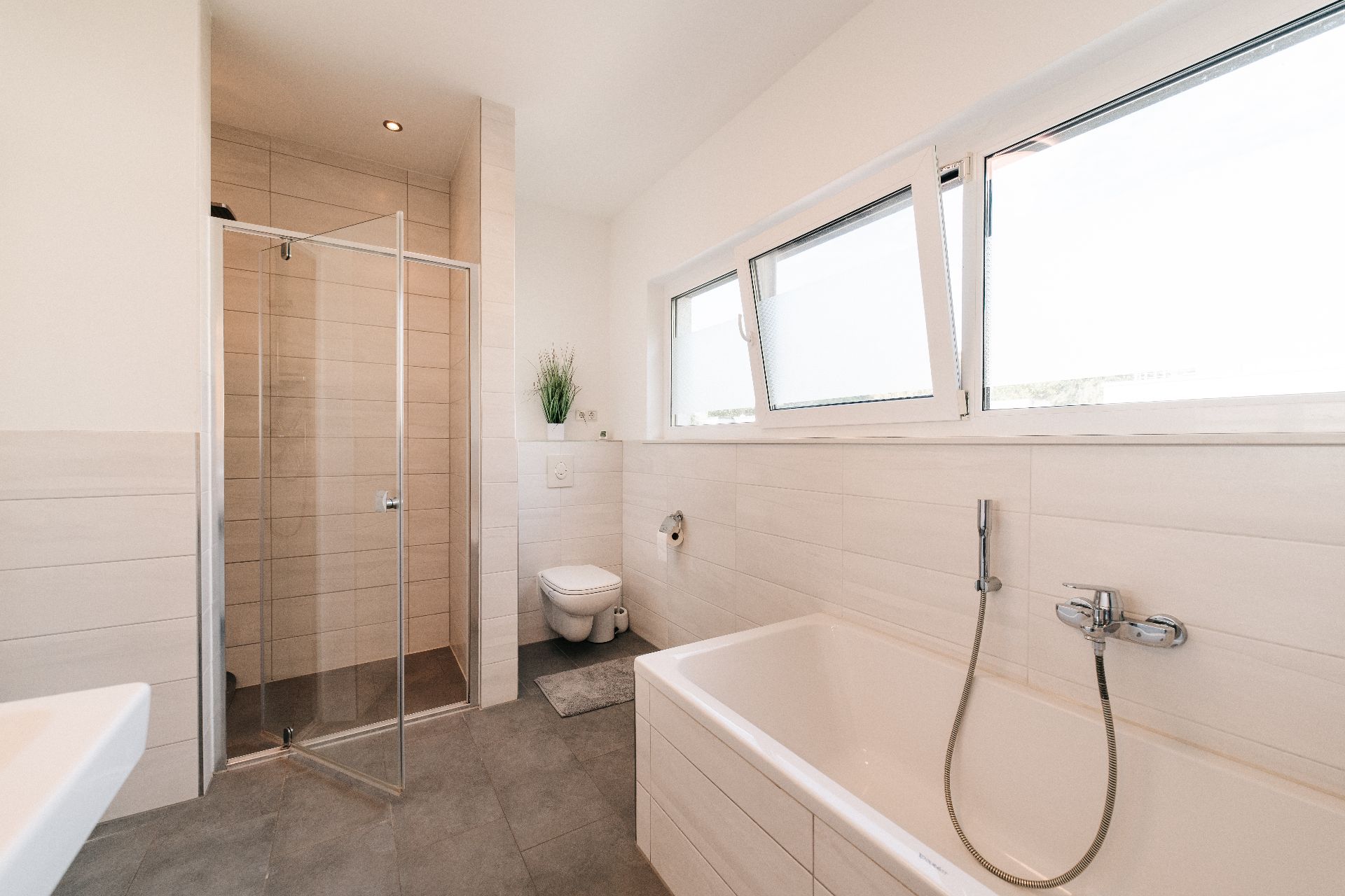 Helles Badezimmer mit großer Badewanne, Dusche, Toilette und drei Fenstern.