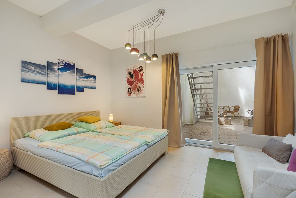 Helles Schlafzimmer mit Doppelbett, Sofa, Wandbildern und Zugang zu einem kleinen Innenhof mit Sitzmöbeln.