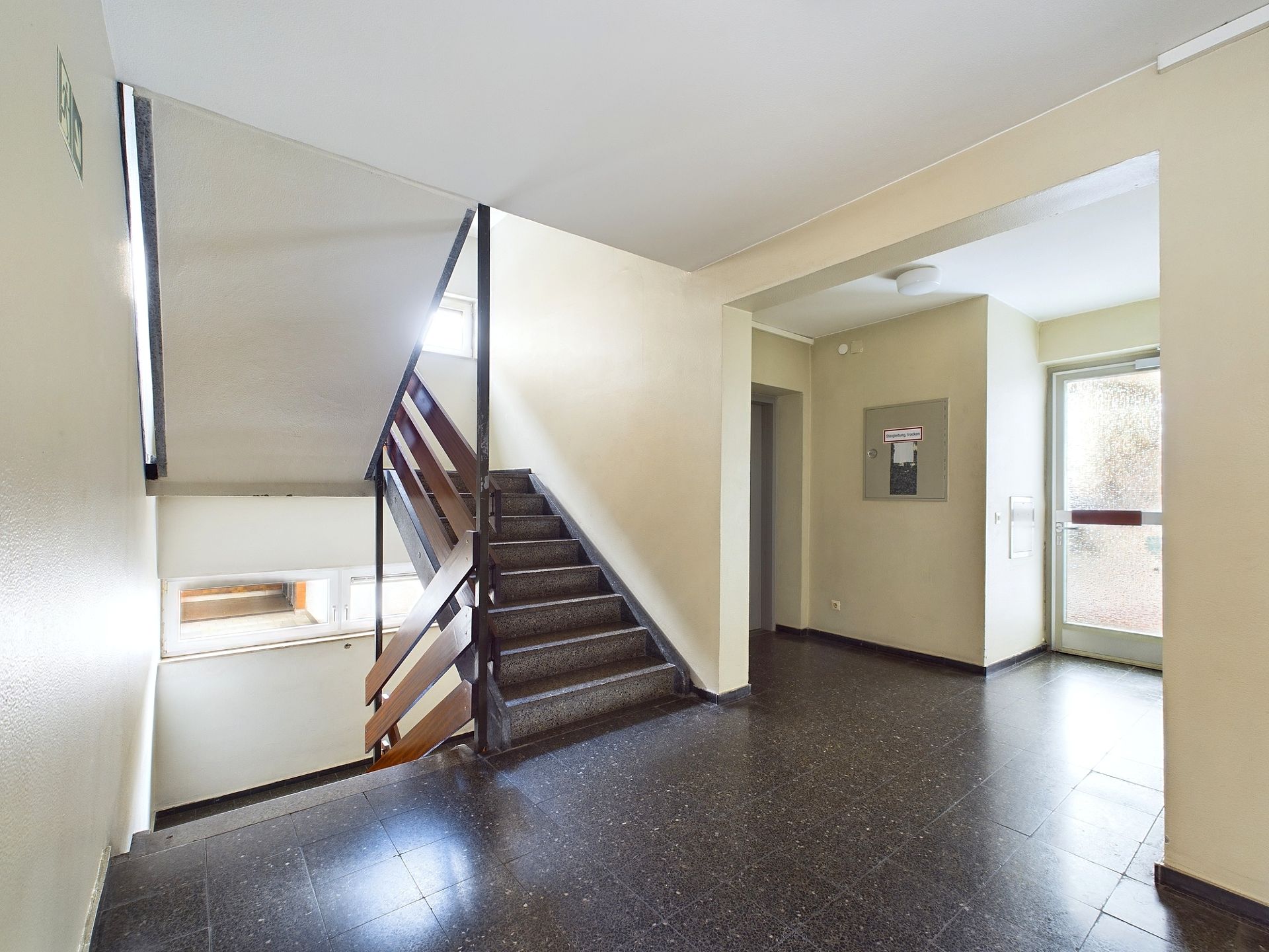 Heller Treppenhausbereich mit Fliesenboden, Treppe und Eingangstür eines Wohngebäudes.