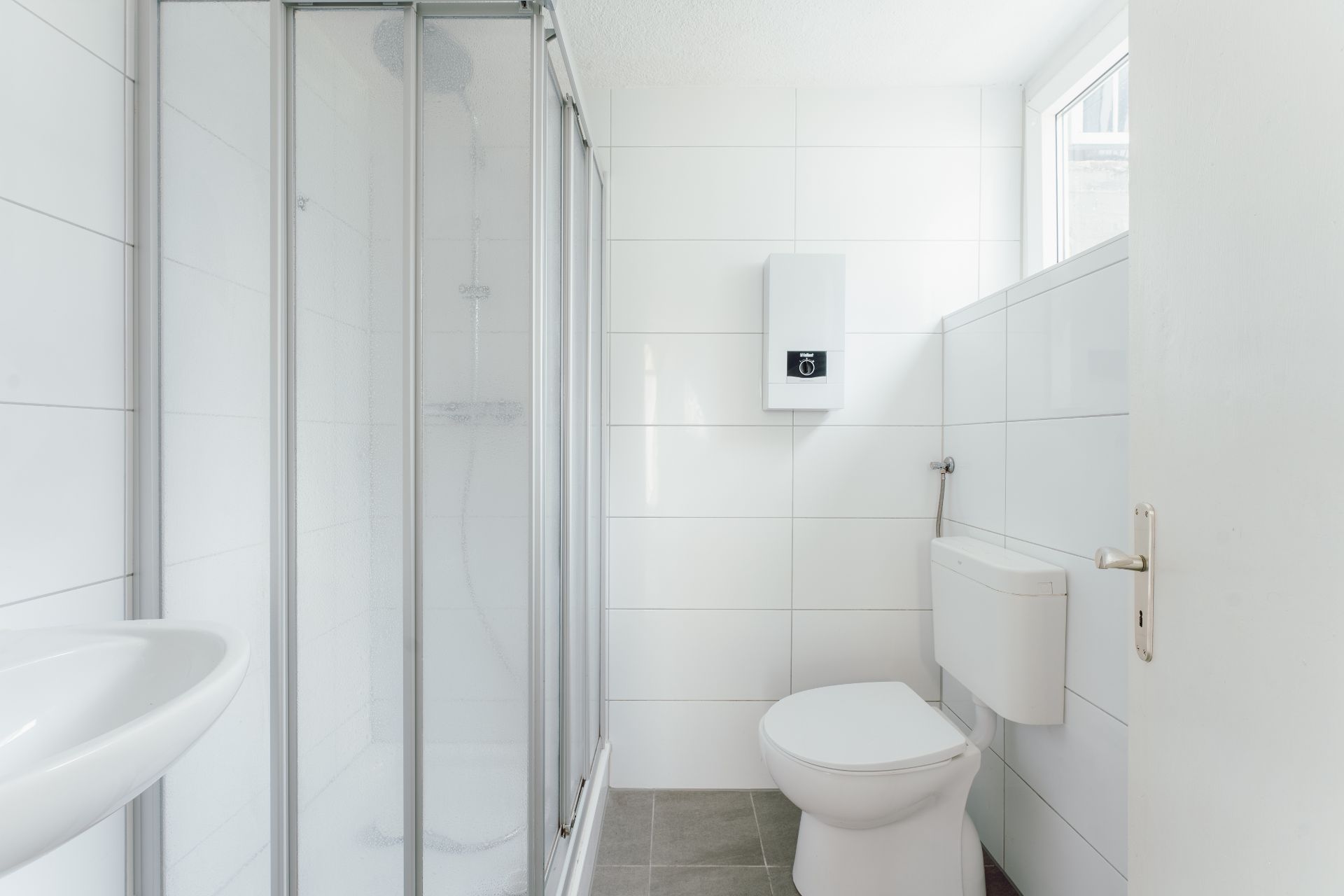 Helles Bad mit Dusche, Toilette und Waschbecken, weiße Fliesen und Fenster.
